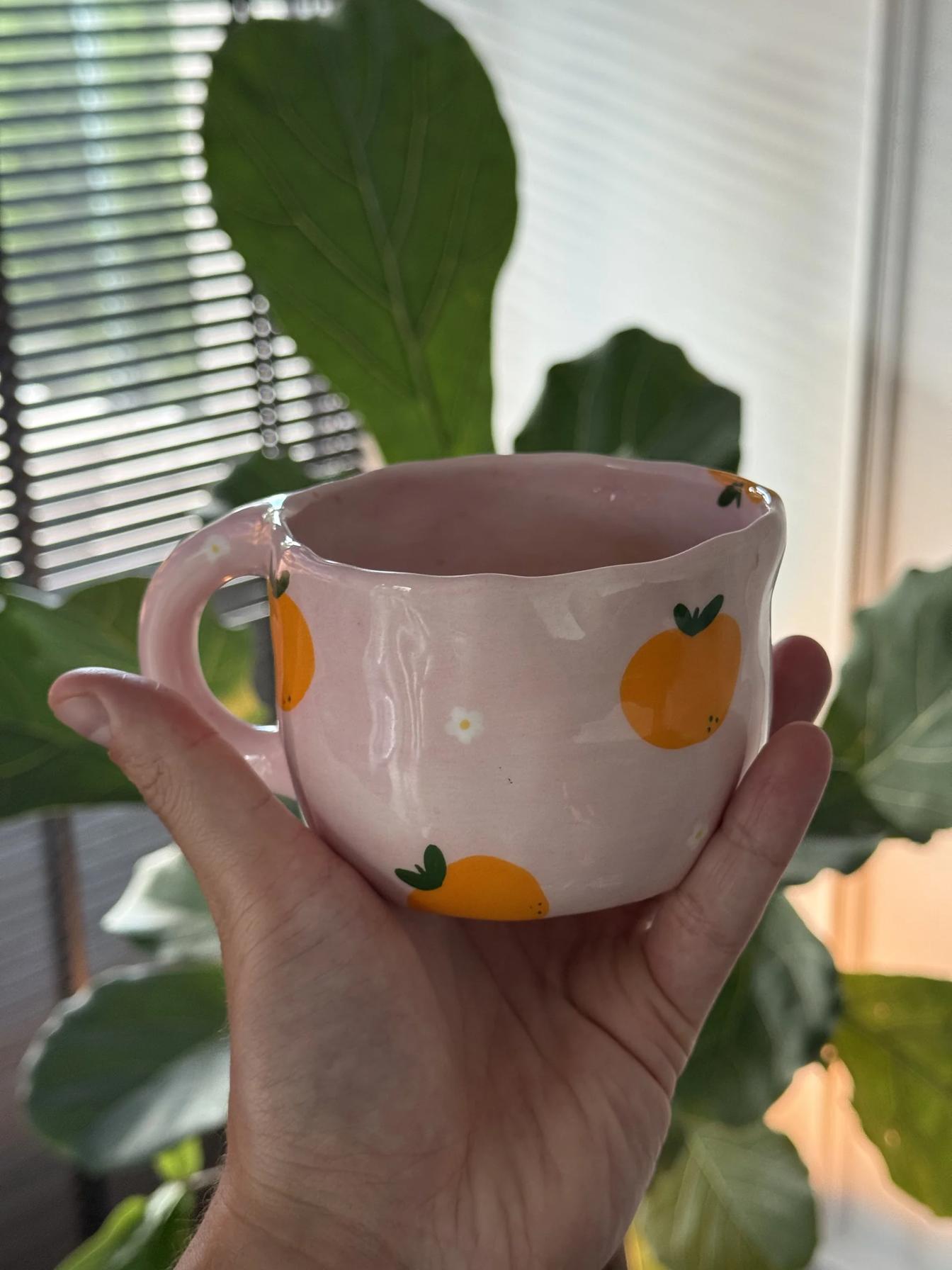 Hand holding a small pink handmade ceramic mug painted with oranges, green leaves, and a tiny white flower