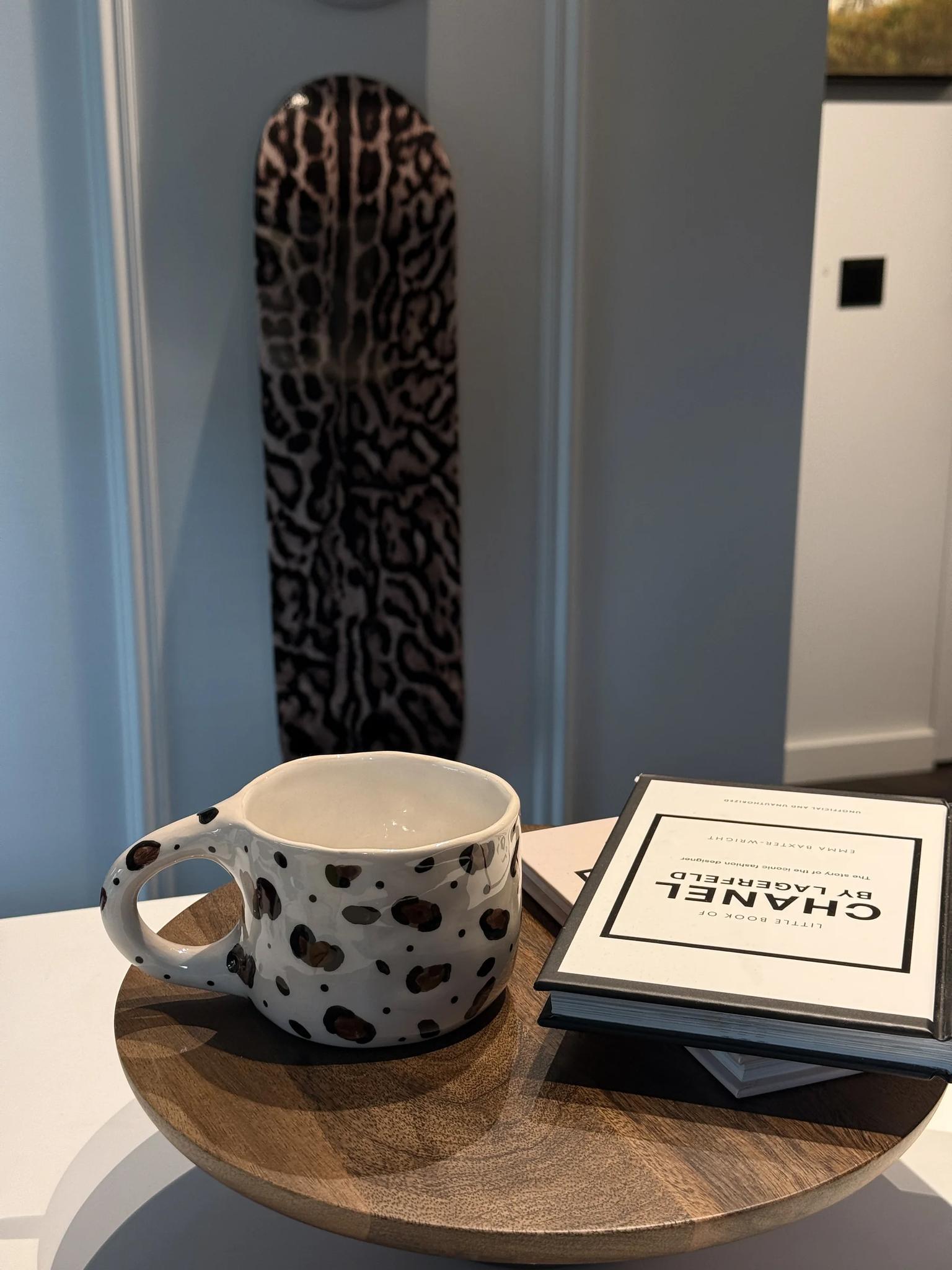 Cream handmade ceramic mug with brown and black leopard spots on a round oak table, with a leopard-print skateboard deck on the wall behind it and a book beside the mug